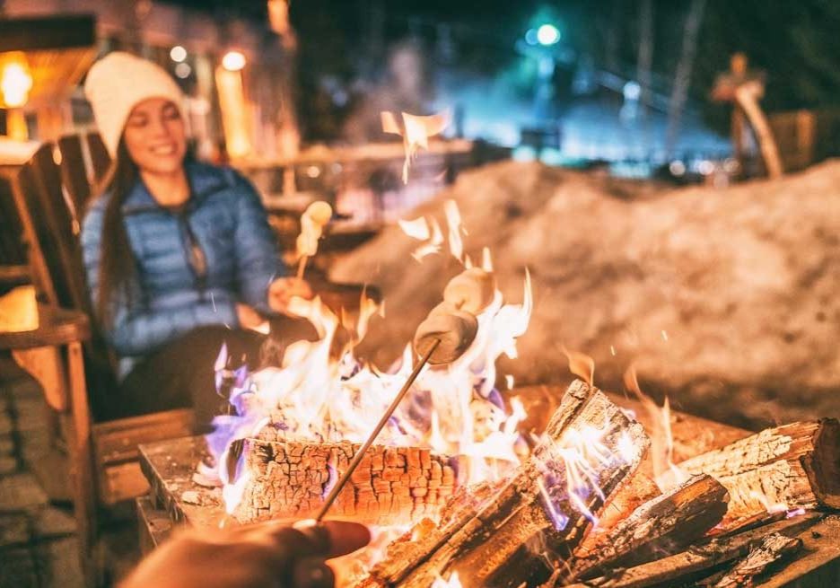 Winter holiday ski resort woman roasting marshmallows in BBQ firepit afterski fun leisure activity with friends. Couple grilling marshmallow stick in fire at Tamarack Resort in Idaho